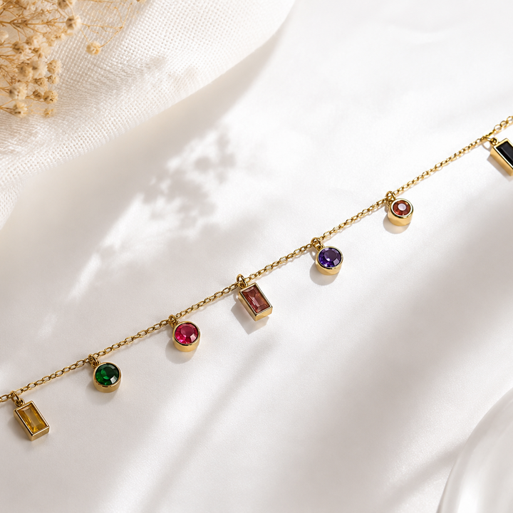 A delicate gold chain bracelet with multiple small colorful gemstone charms and rectangular pendant charms, displayed on a soft white fabric background with dried flowers in the corner.