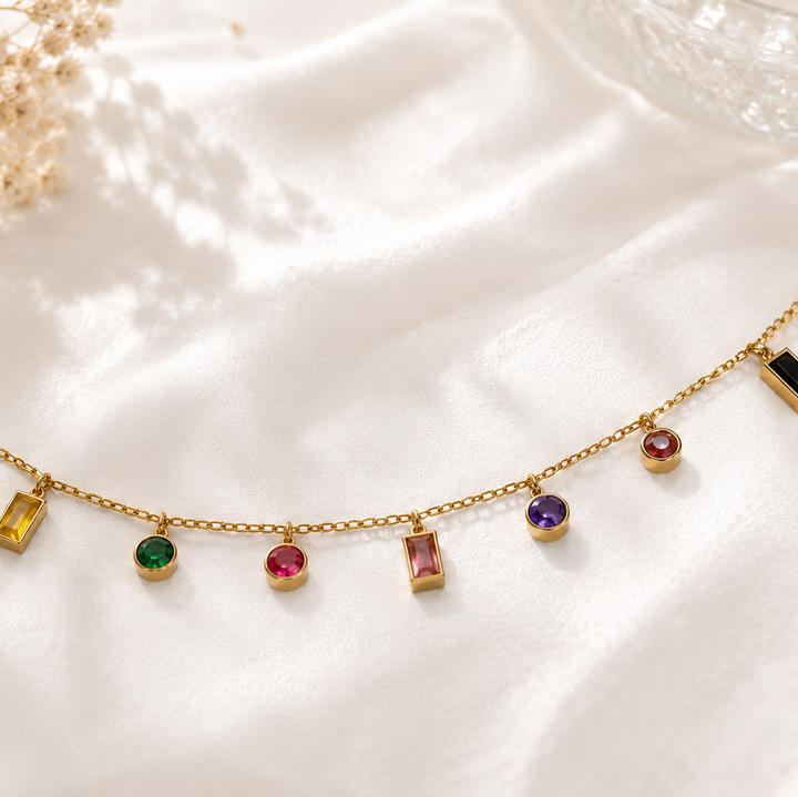 A gold chain BRACELET with multiple small, colorful gemstone charms in various shapes, displayed on a draped white satin fabric.