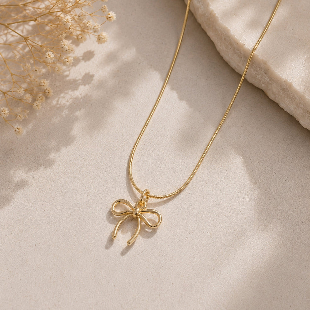Gold necklace with a bow pendant on a light beige surface with dried flowers.