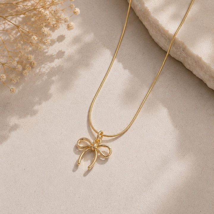 Gold necklace with a bow pendant on a light beige surface with dried flowers.