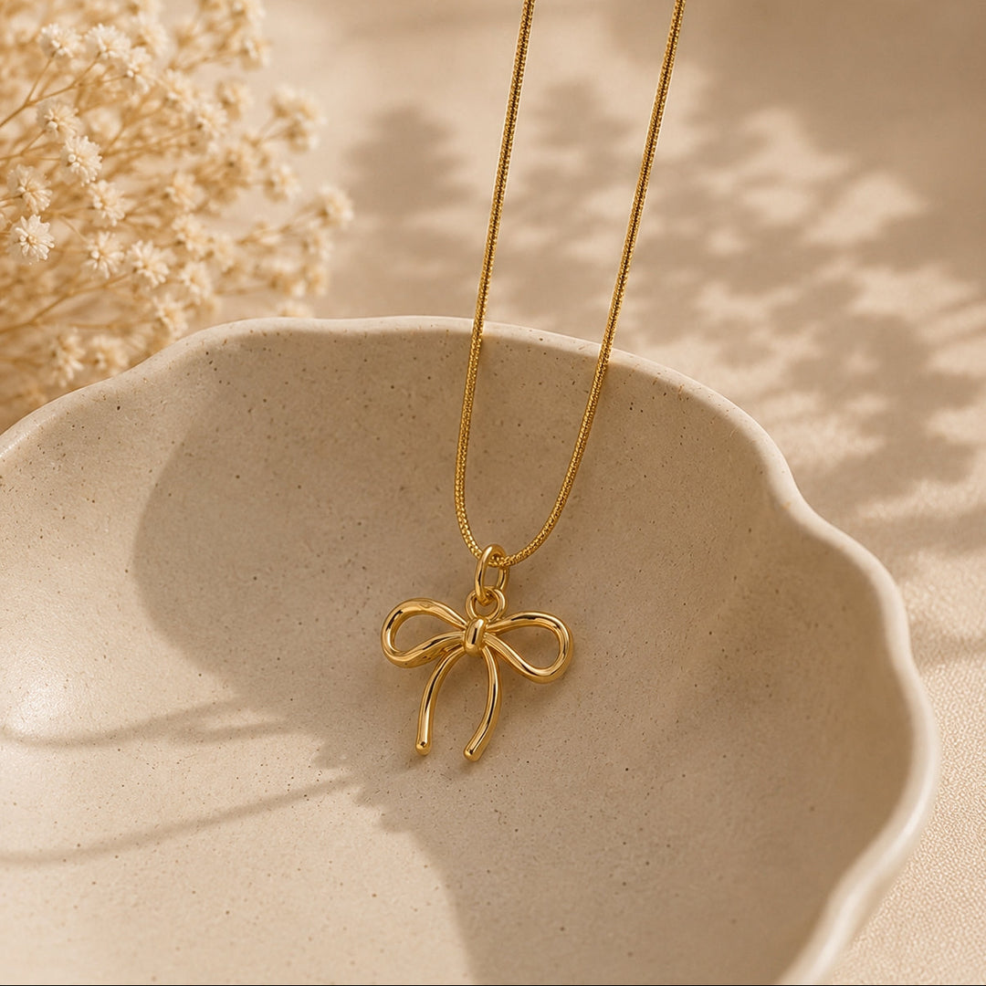 Gold bow-shaped pendant necklace on a beige dish with dried flowers.