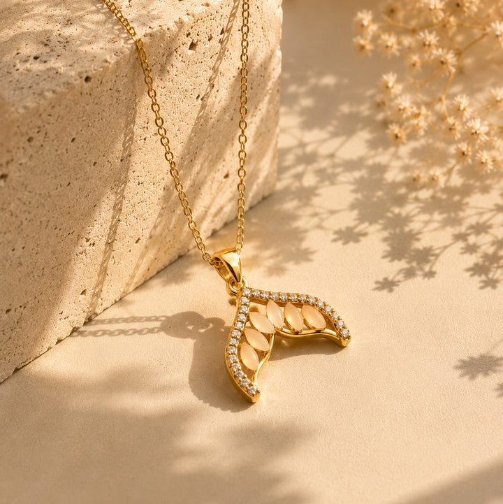 Gold necklace with a leaf-shaped pendant on a stone surface with dried flowers.