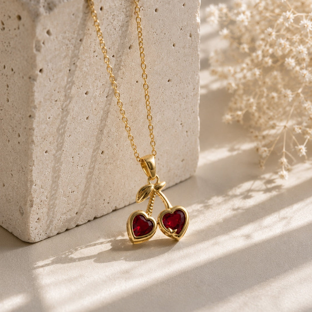 Gold necklace with red heart pendants on a textured surface with dried flowers.