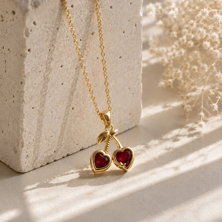 Gold necklace with red heart pendants on a textured surface with dried flowers.
