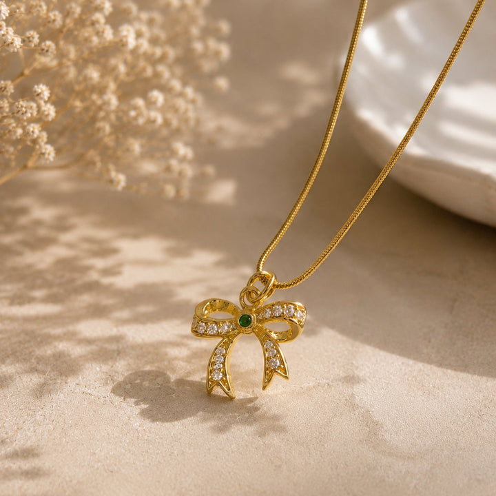 Gold necklace with a bow-shaped pendant on a beige surface with dried flowers.