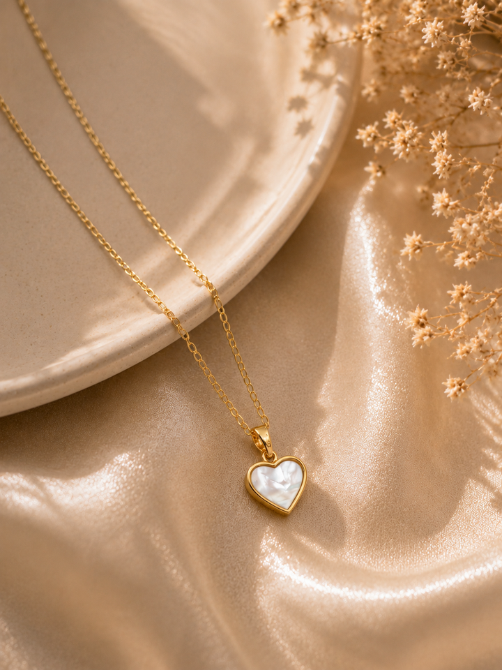 Gold necklace with a heart-shaped pendant on a beige surface with dried flowers.