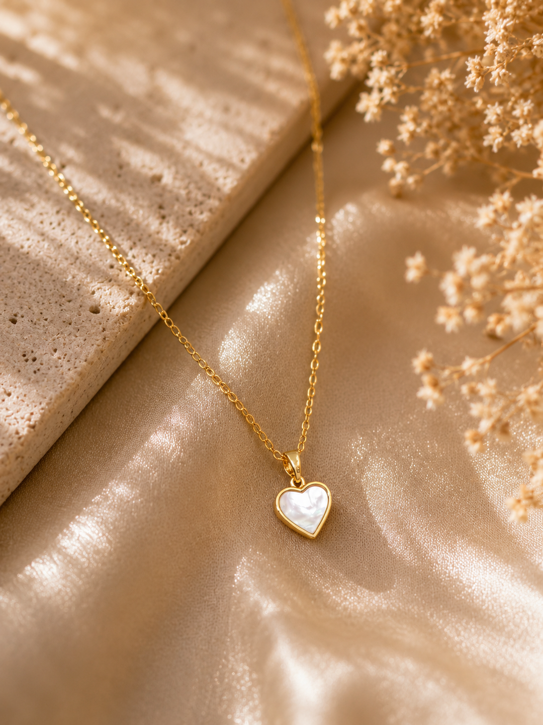 Gold necklace with a heart-shaped pendant on a beige surface with dried flowers.