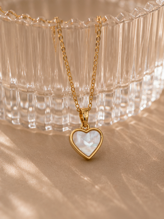 Gold heart-shaped pendant necklace on a beige surface with a clear glass dish in the background