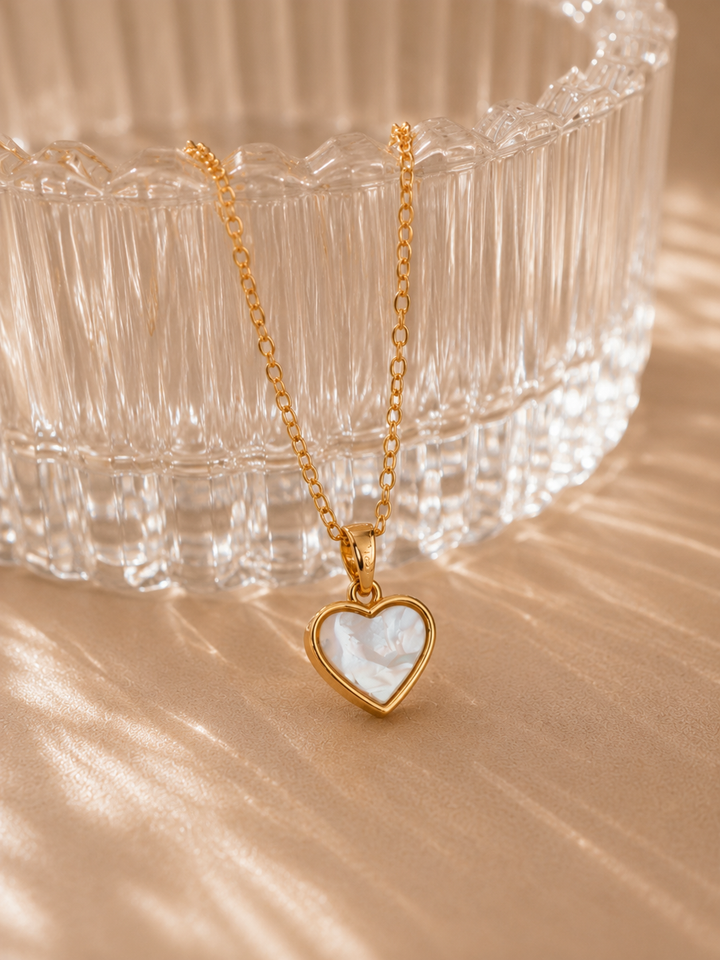 Gold necklace with a heart-shaped pendant on a beige surface with a glass bowl in the background