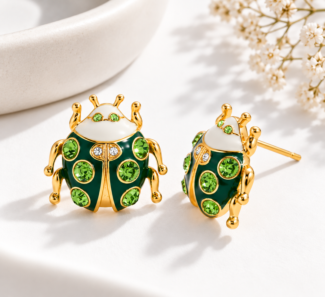 A pair of gold-plated ladybug stud earrings with green crystal accents and a white enamel head, displayed on a clean white surface with soft shadows and small dried flowers in the background.
