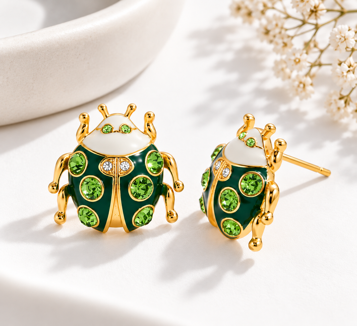 A pair of gold-plated ladybug stud earrings with green crystal accents and a white enamel head, displayed on a clean white surface with soft shadows and small dried flowers in the background.