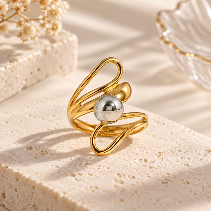 A gold-toned abstract ring with a central polished silver bead, displayed on a textured beige stone slab with soft shadows and decorative props in the background.