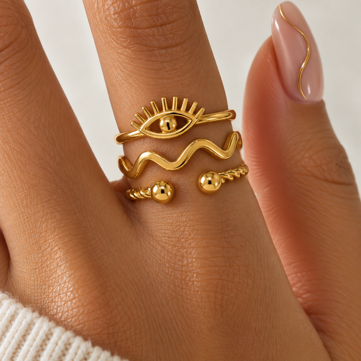 A close-up of a hand wearing three stacked gold rings: an eye motif ring, a wavy band ring, and a beaded open-band ring. The hand has pale pink manicured nails and is wearing a white knit sweater.