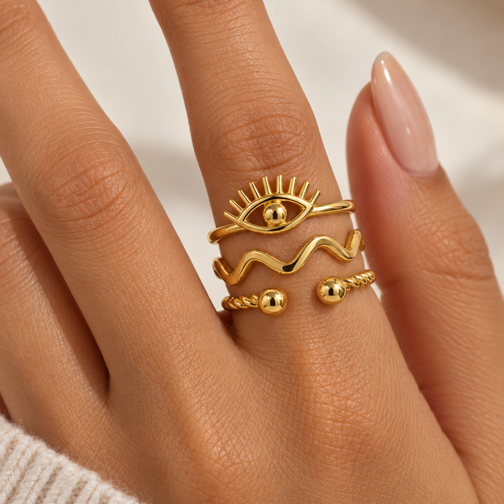A close-up of a hand wearing three stacked gold rings with bohemian designs, including a sun, wavy lines, and small beads, paired with a cream knit sweater.