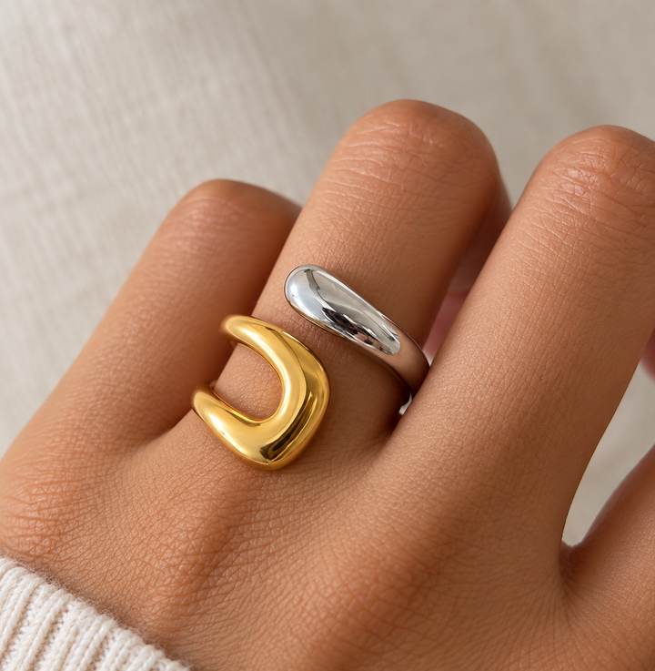 A close-up of a hand wearing two abstract open rings, one in polished gold and one in polished silver, against a soft beige textured background.