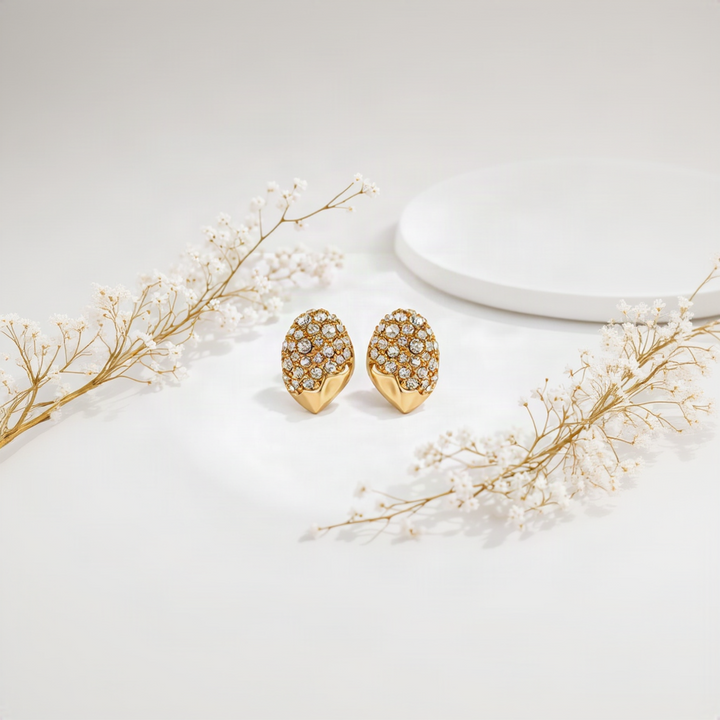 A pair of gold-toned stud earrings with a textured, rounded top covered in clear pavé rhinestones, set against a soft white background with subtle floral props.