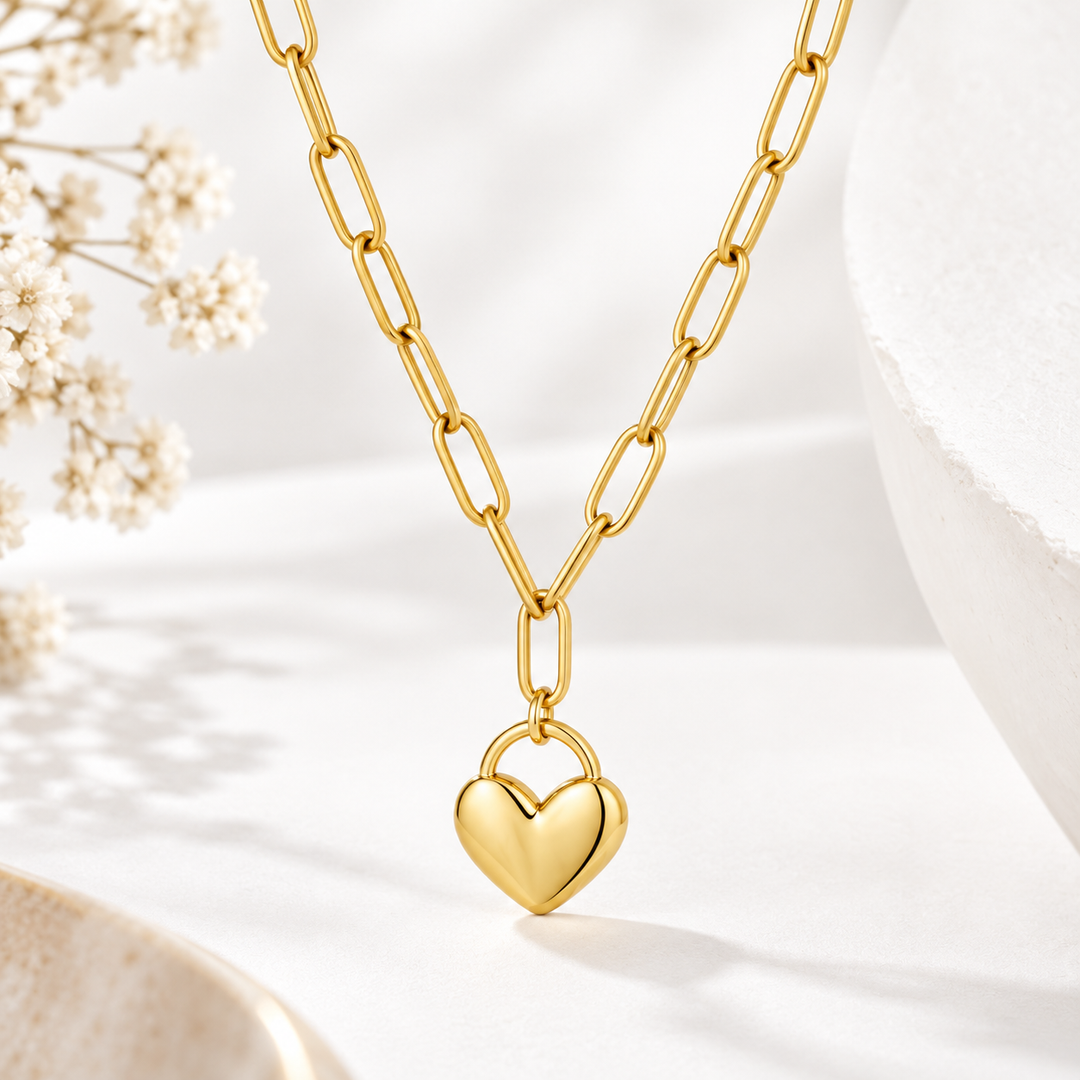 A gold paperclip chain necklace with a polished gold heart pendant, displayed on a clean white surface with soft shadows and decorative dried flowers.