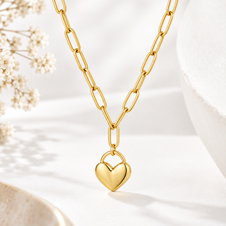 A gold paperclip chain necklace with a polished gold heart pendant, displayed on a clean white surface with soft shadows and decorative dried flowers.