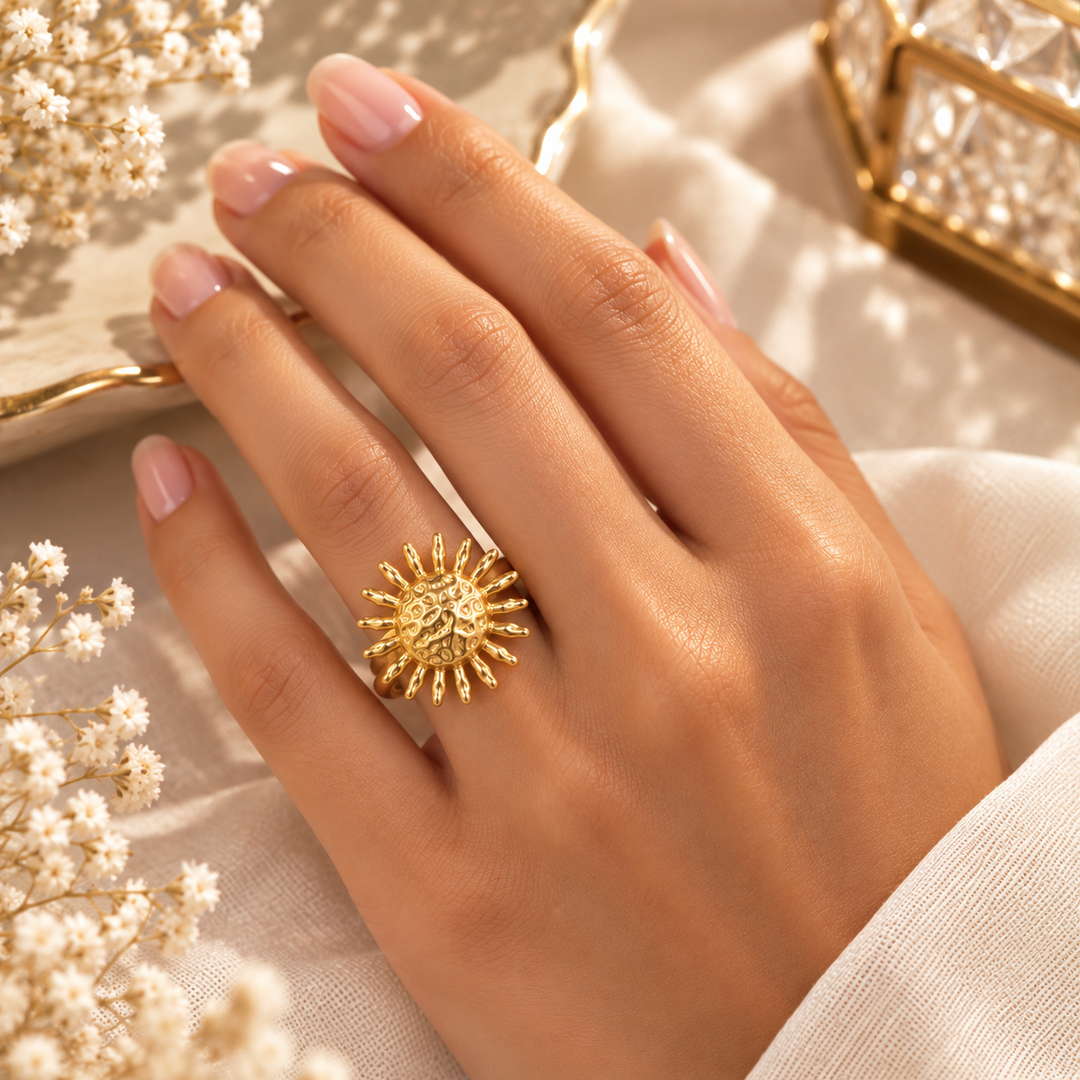 A close-up of a hand wearing a textured gold sunburst ring, styled with neutral cream fabric and small white flowers.