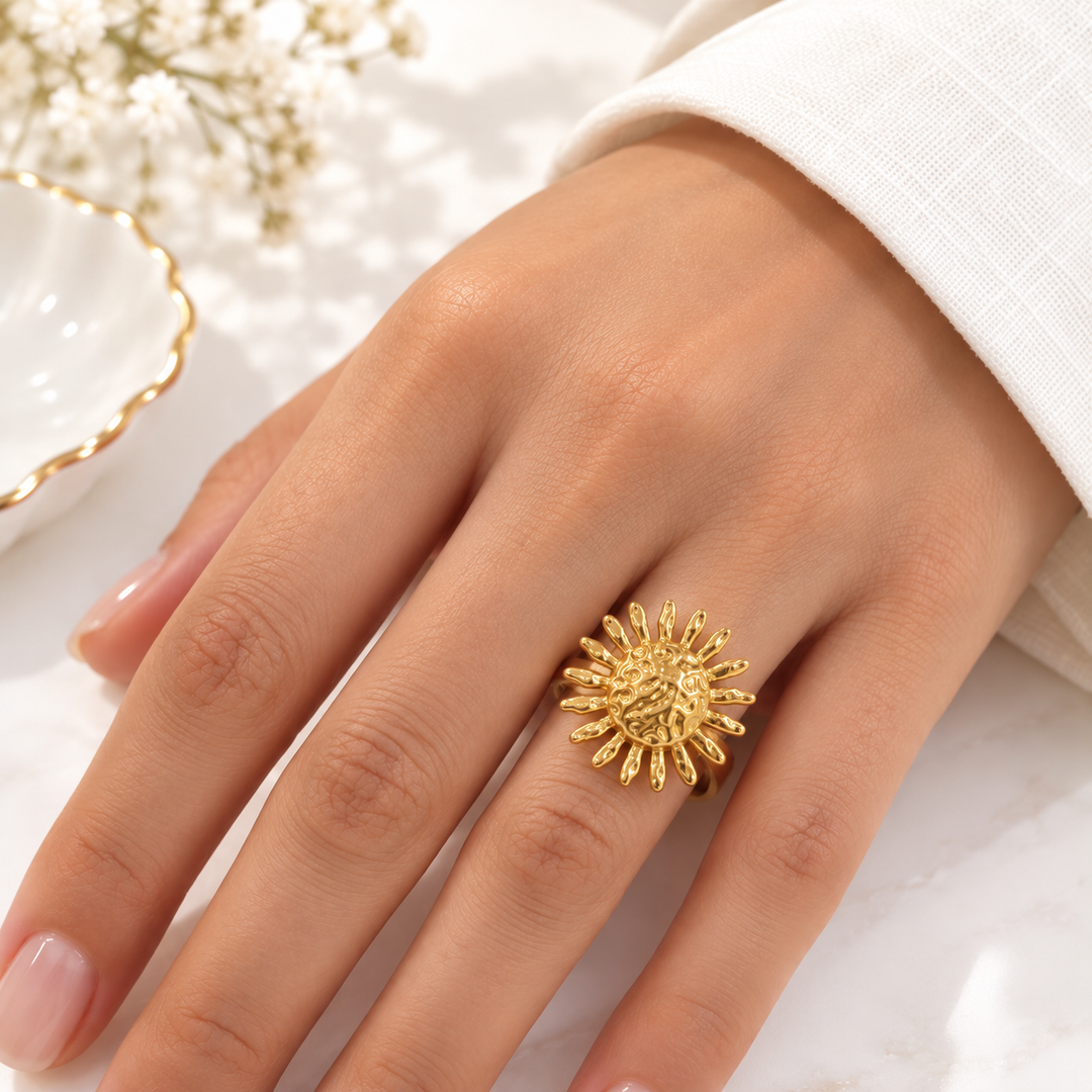 A close-up of a hand wearing a textured, sunburst-shaped gold ring, styled with a white sleeve and delicate white flowers in the background.