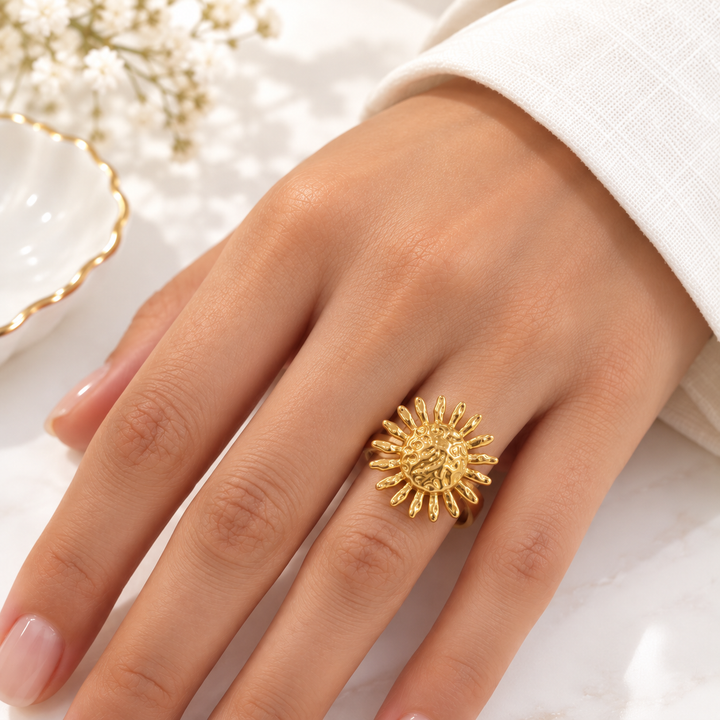 A close-up of a hand wearing a textured, sunburst-shaped gold ring, styled with a white sleeve and delicate white flowers in the background.