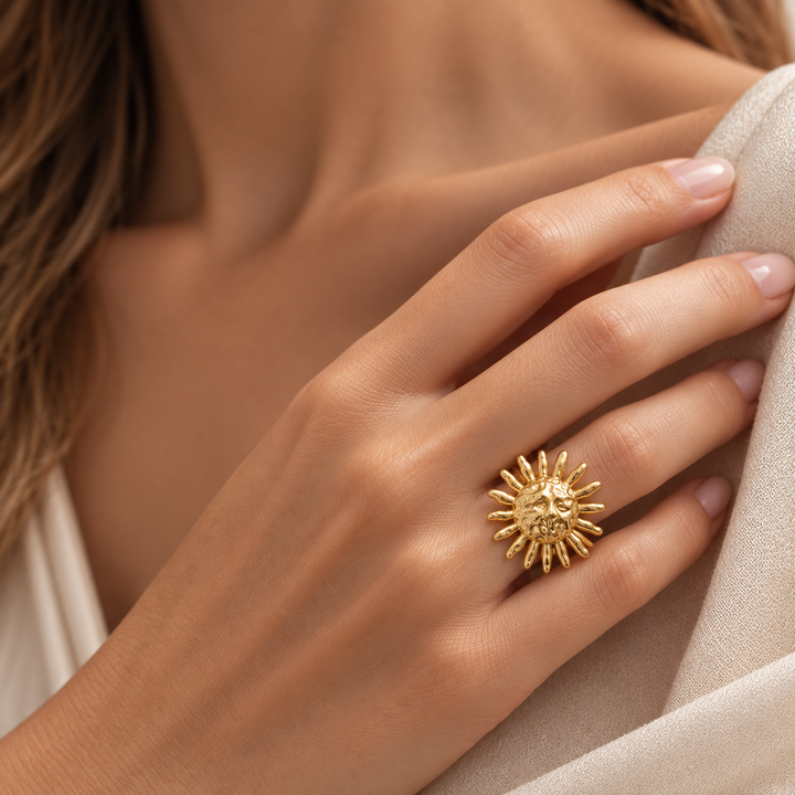 A close-up of a hand wearing a textured gold sunburst ring, resting against a neutral beige fabric background.