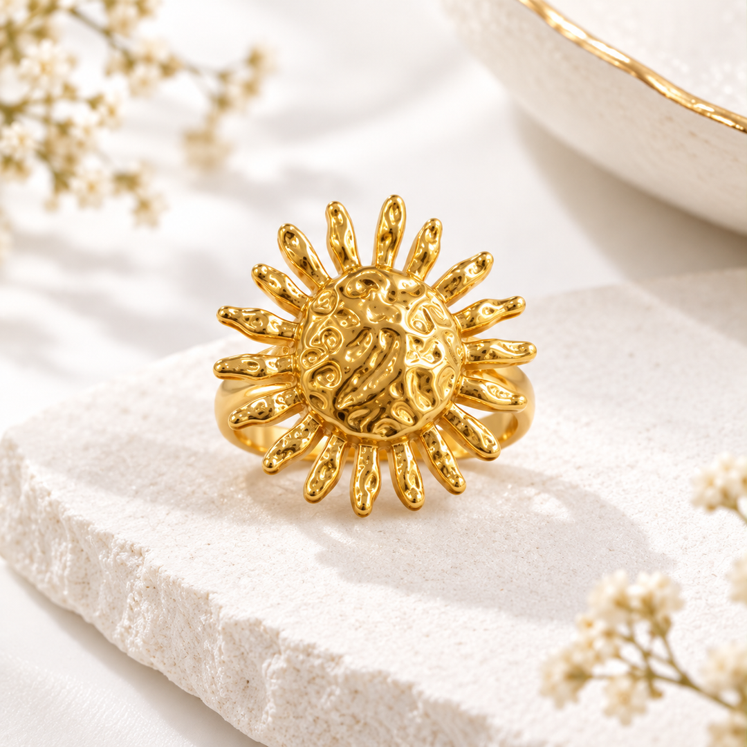 A gold sun-shaped ring with textured rays and a central floral motif, displayed on a white stone surface with delicate baby's breath flowers and a white ceramic bowl in the background.