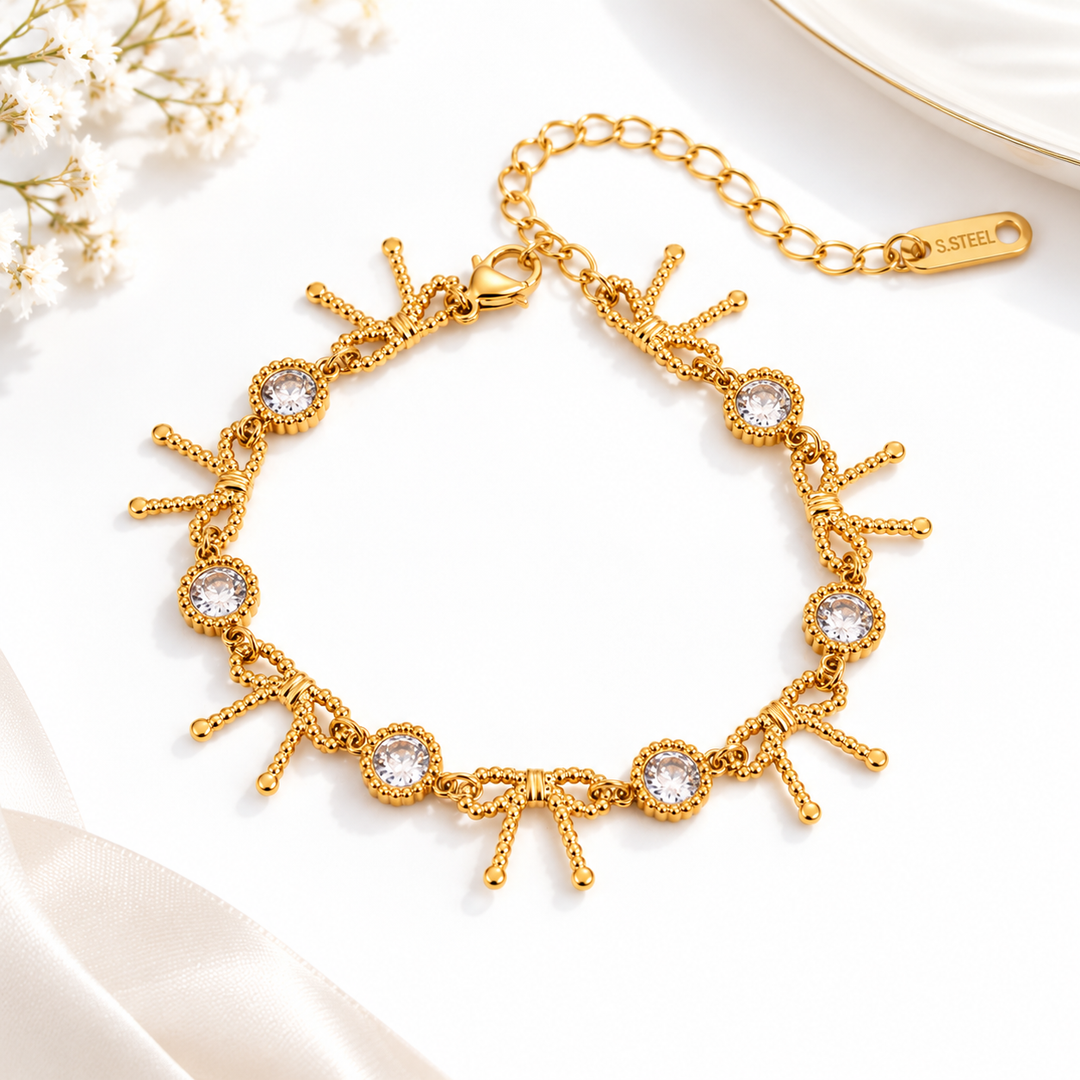 A gold-tone chain bracelet with decorative bow and circular crystal accents, displayed on a clean white surface with soft floral and satin props.