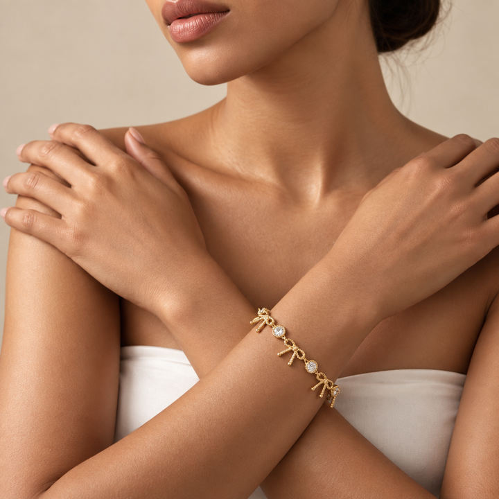 A close-up of a model wearing a delicate gold chain bracelet with small bow charms and pearl accents on her wrist, paired with a white strapless top against a neutral beige background.