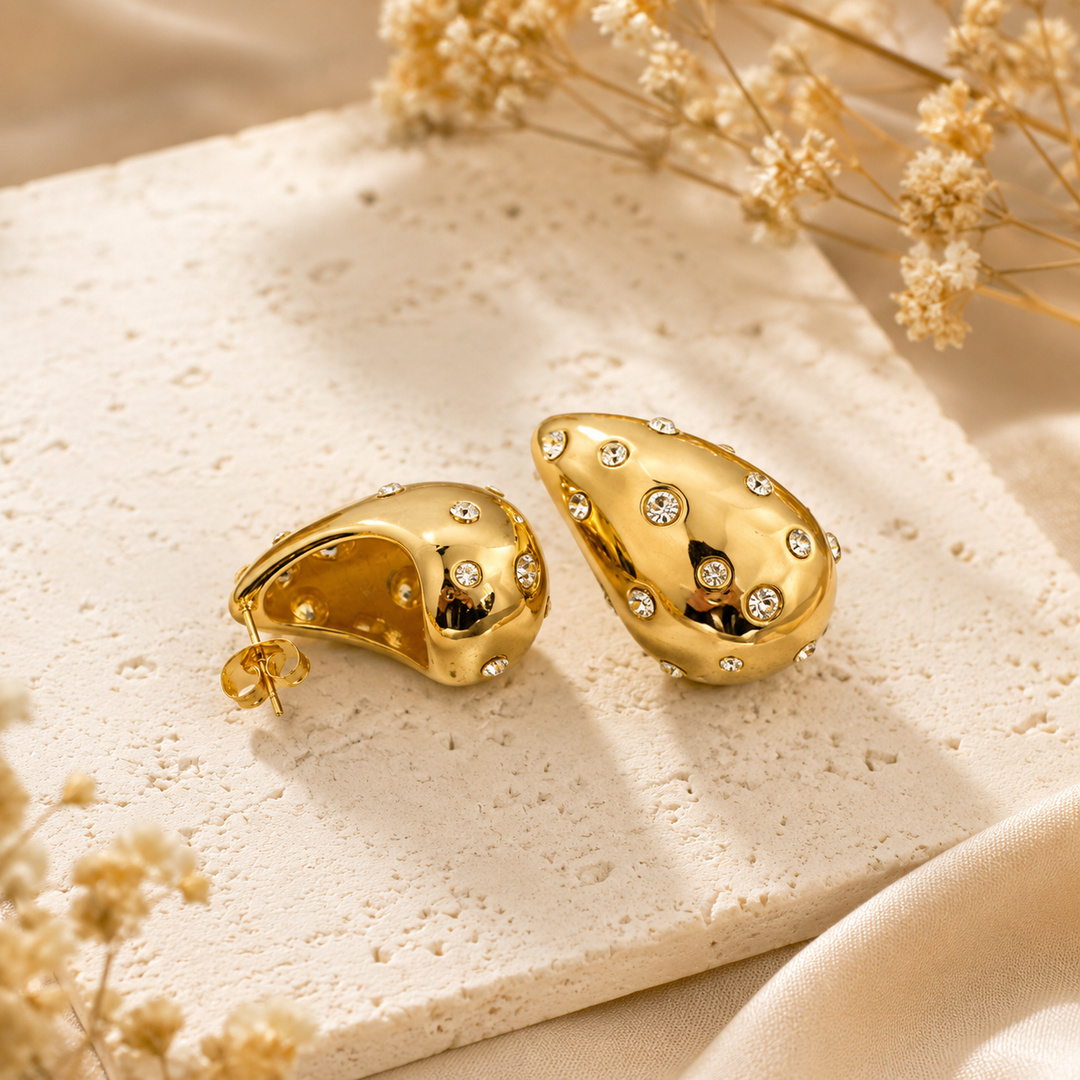 A pair of gold teardrop-shaped earrings adorned with small clear rhinestones, displayed on a textured beige stone slab with dried flowers.