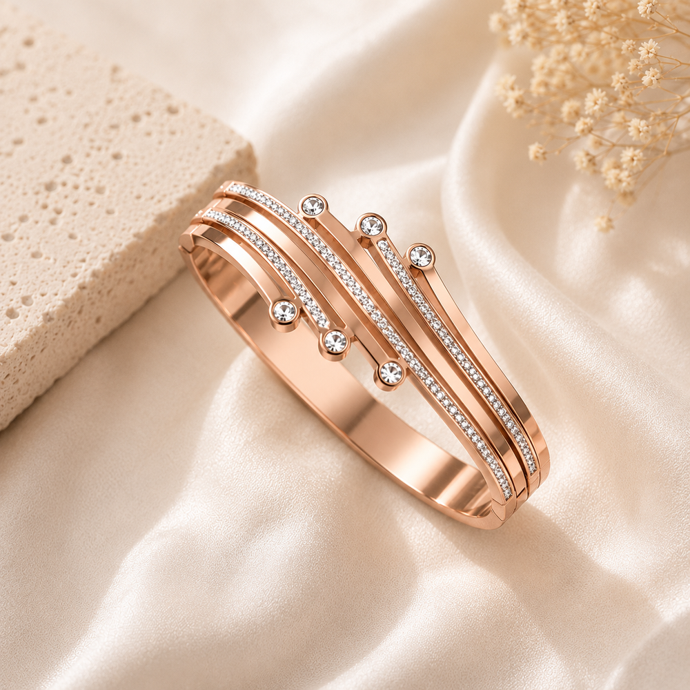 A rose gold bangle bracelet with multiple parallel bands, some adorned with small clear stones, displayed on a draped cream satin fabric with a textured stone and dried flowers as props.