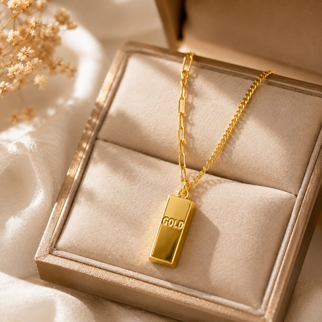 A gold bar pendant necklace resting in an open beige jewelry box with a cream velvet interior. The pendant is engraved with the word 'GOLD' and hangs from a gold chain.