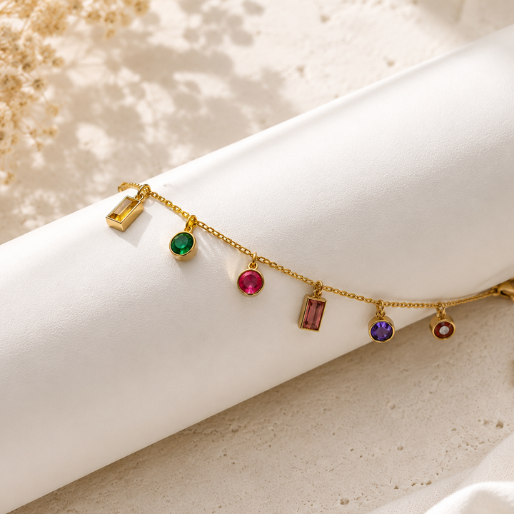 A delicate gold chain bracelet with multiple colorful gemstone charms, displayed on a white cylindrical stand against a light beige textured background.