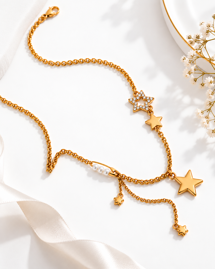 A gold-toned chain necklace with multiple star charms, some adorned with small clear rhinestones, a pearl bead, and dangling star accents, styled on a white surface with soft satin ribbon and dried flowers.