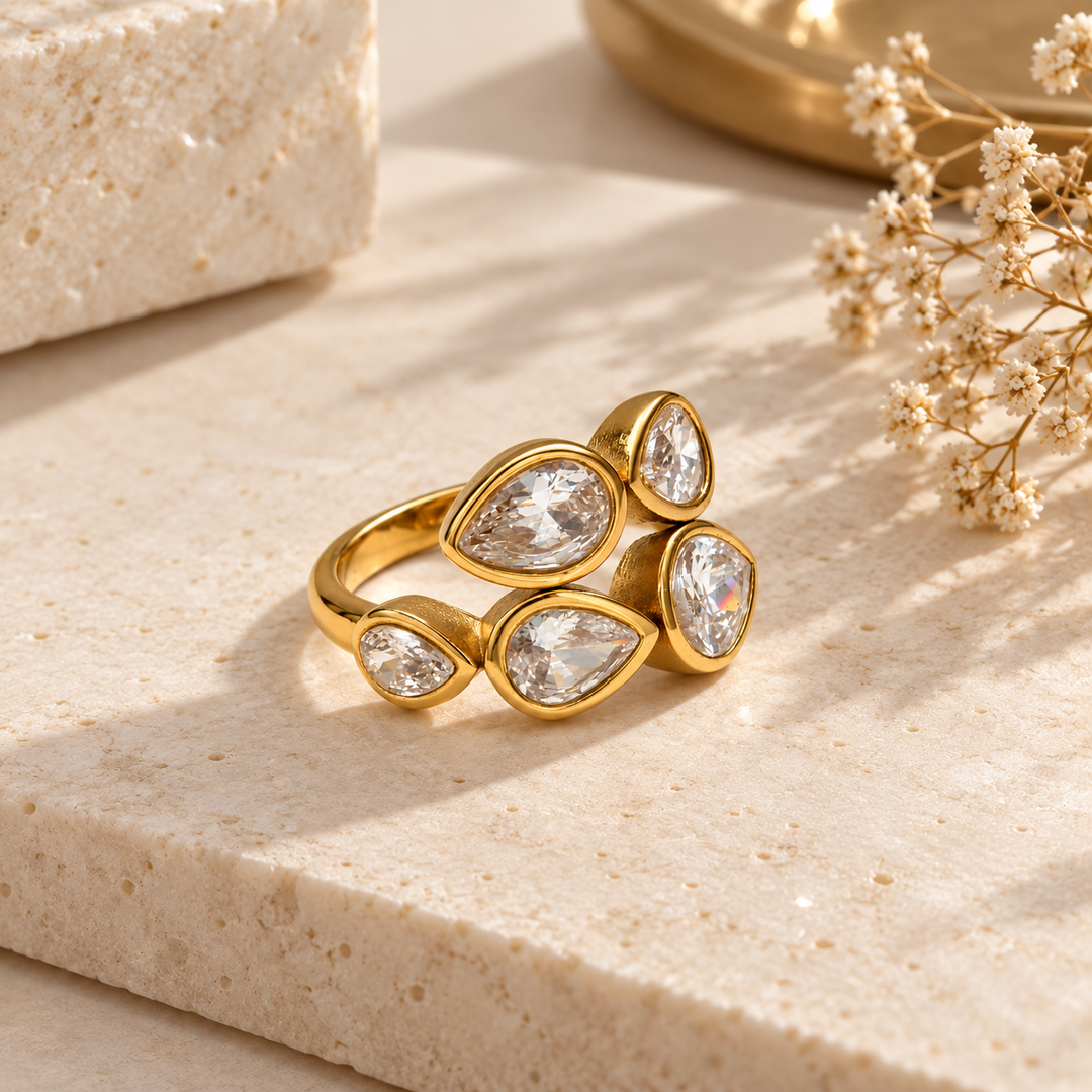 A gold ring with five teardrop-shaped clear gemstones, styled on a beige stone slab with dried flowers and a gold tray in the background.
