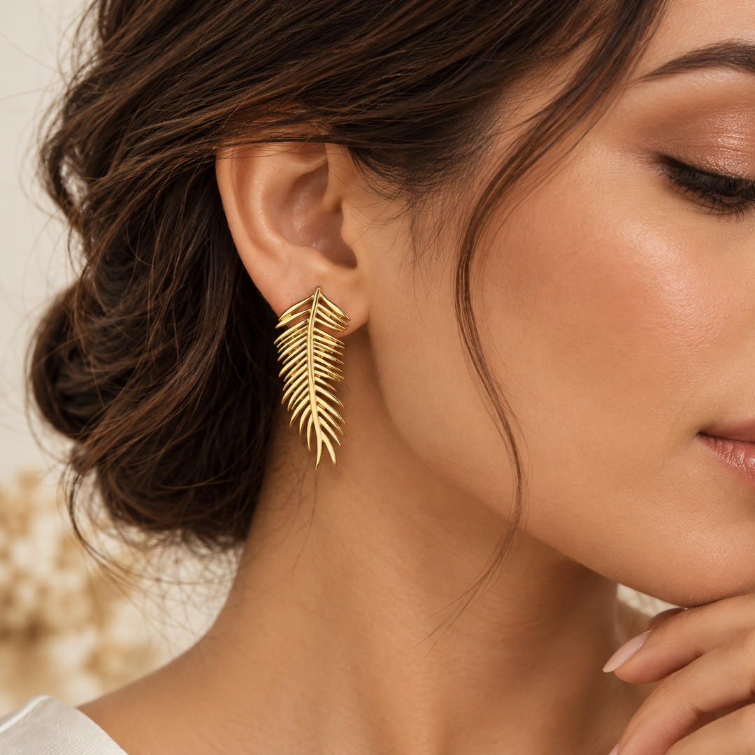 A close-up of a woman wearing a single gold leaf-shaped dangle earring. The earring has a textured, layered leaf design with fine parallel lines.