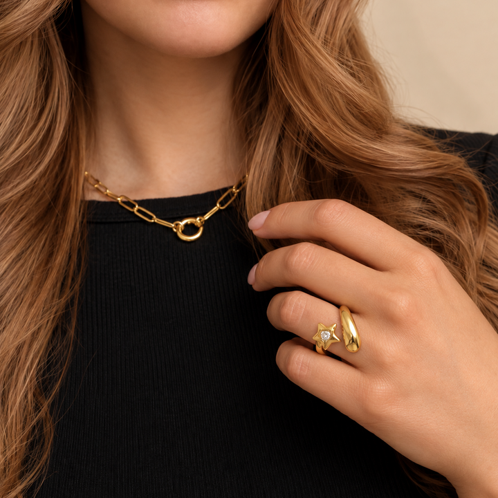 A close-up lifestyle shot of a woman wearing a gold chain necklace and a gold ring with a clear gemstone, styled with a black ribbed top against a soft beige background.