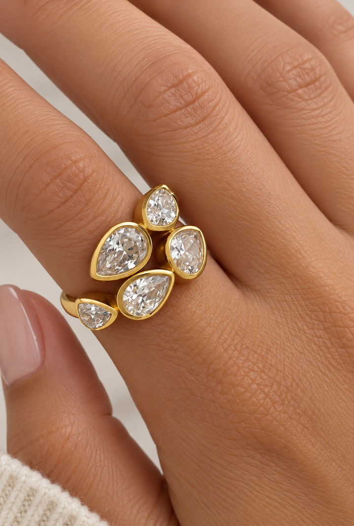 A close-up of a hand wearing a gold ring with five pear-cut clear gemstones in a clustered leaf-like design.