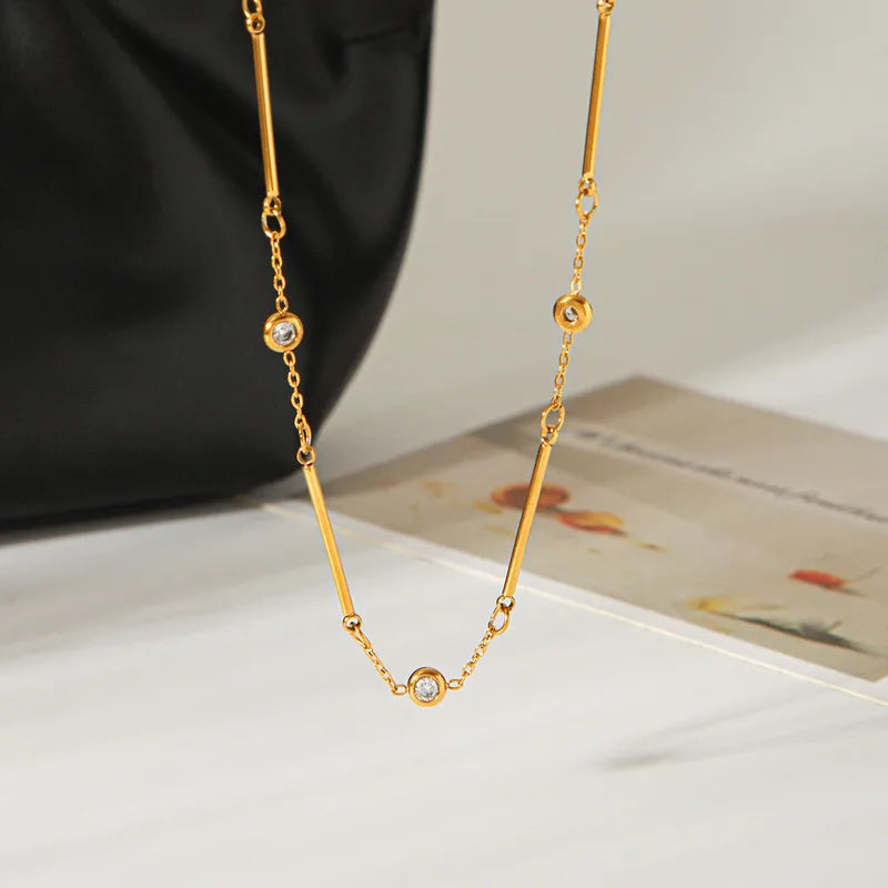 A gold chain necklace with three small round bezel-set clear stones, draped over a white surface with a black bag and a printed card in the background.