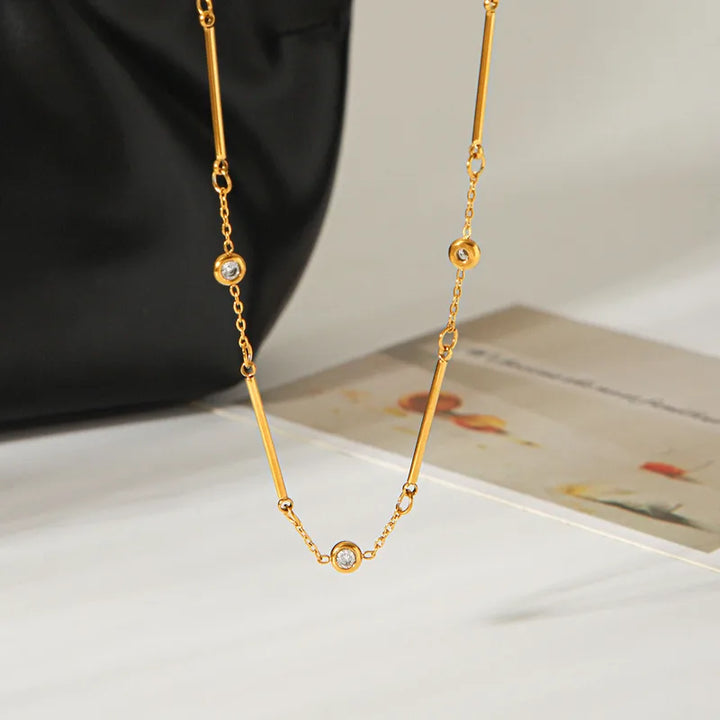 A gold chain necklace with three small round bezel-set clear stones, draped over a white surface with a black bag and a printed card in the background.