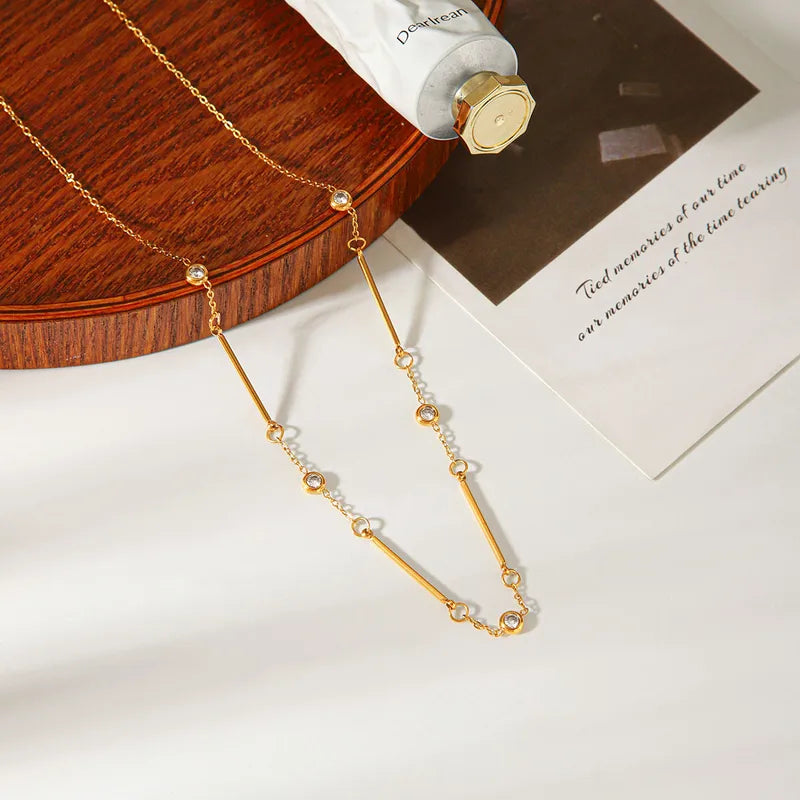 A delicate gold necklace with a long central bar, small bead accents, and clear crystal details, styled on a white surface with a wooden tray, a tube, and a printed card.