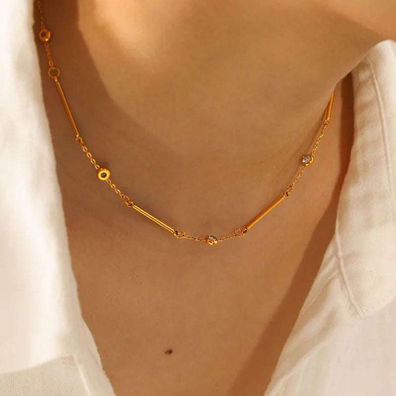 A close-up of a delicate gold necklace with geometric links and small clear stones, worn on a model&