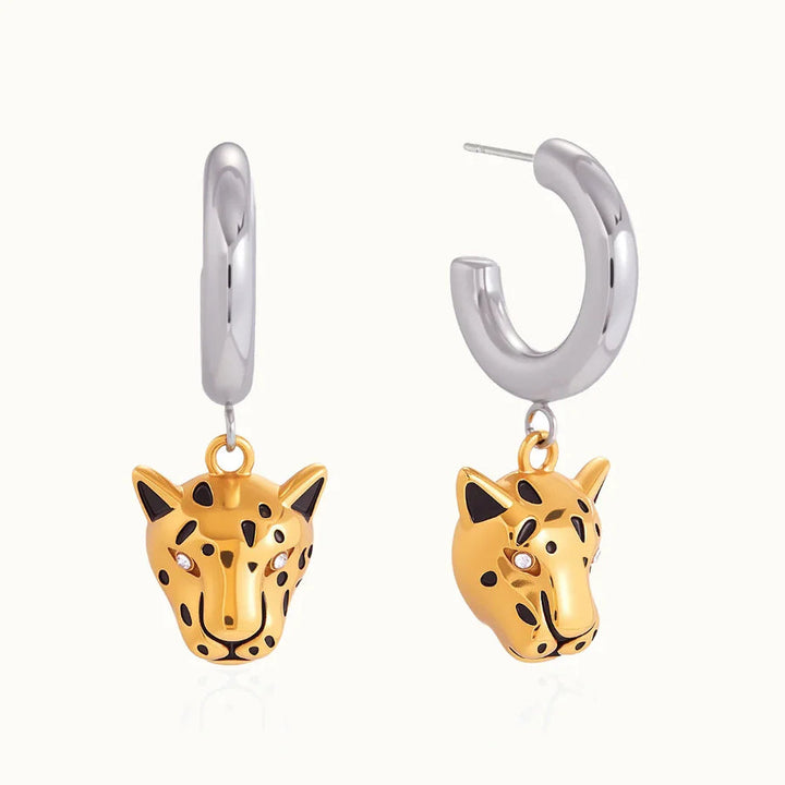 A pair of gold and silver huggie hoop earrings with dangling leopard head charms, shown on a clean off-white background.