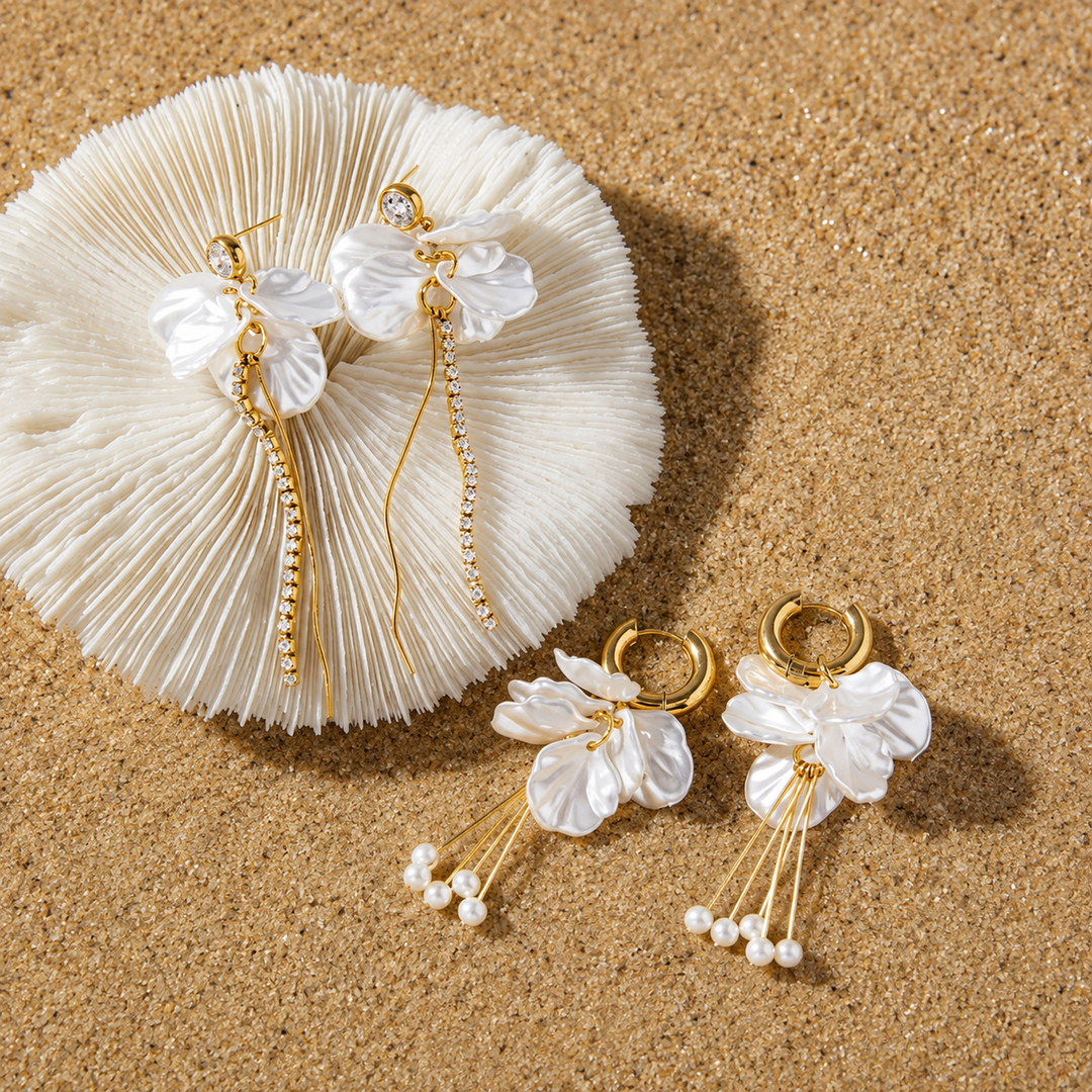 A pair of gold-tone dangle earrings with white flower-shaped charms and small pearl-like beads, displayed on a sandy surface next to a white textured fan.