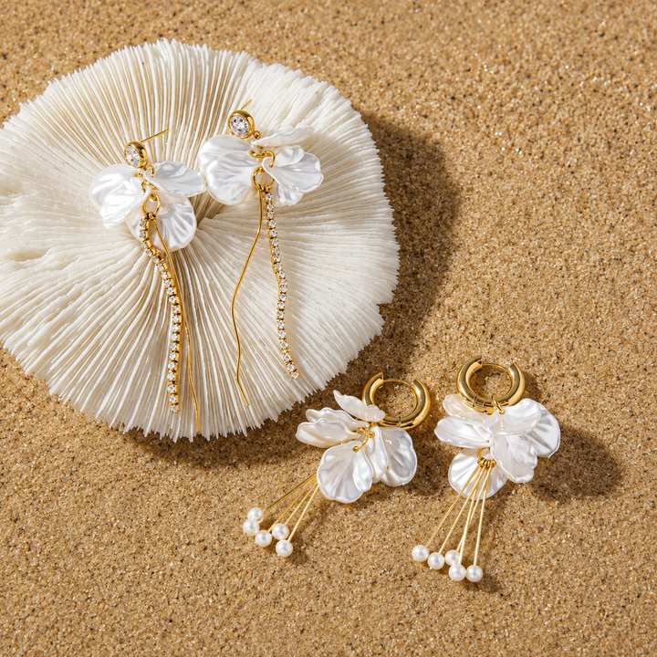 A pair of gold-tone dangle earrings with white flower-shaped charms and small pearl-like beads, displayed on a sandy surface next to a white textured fan.