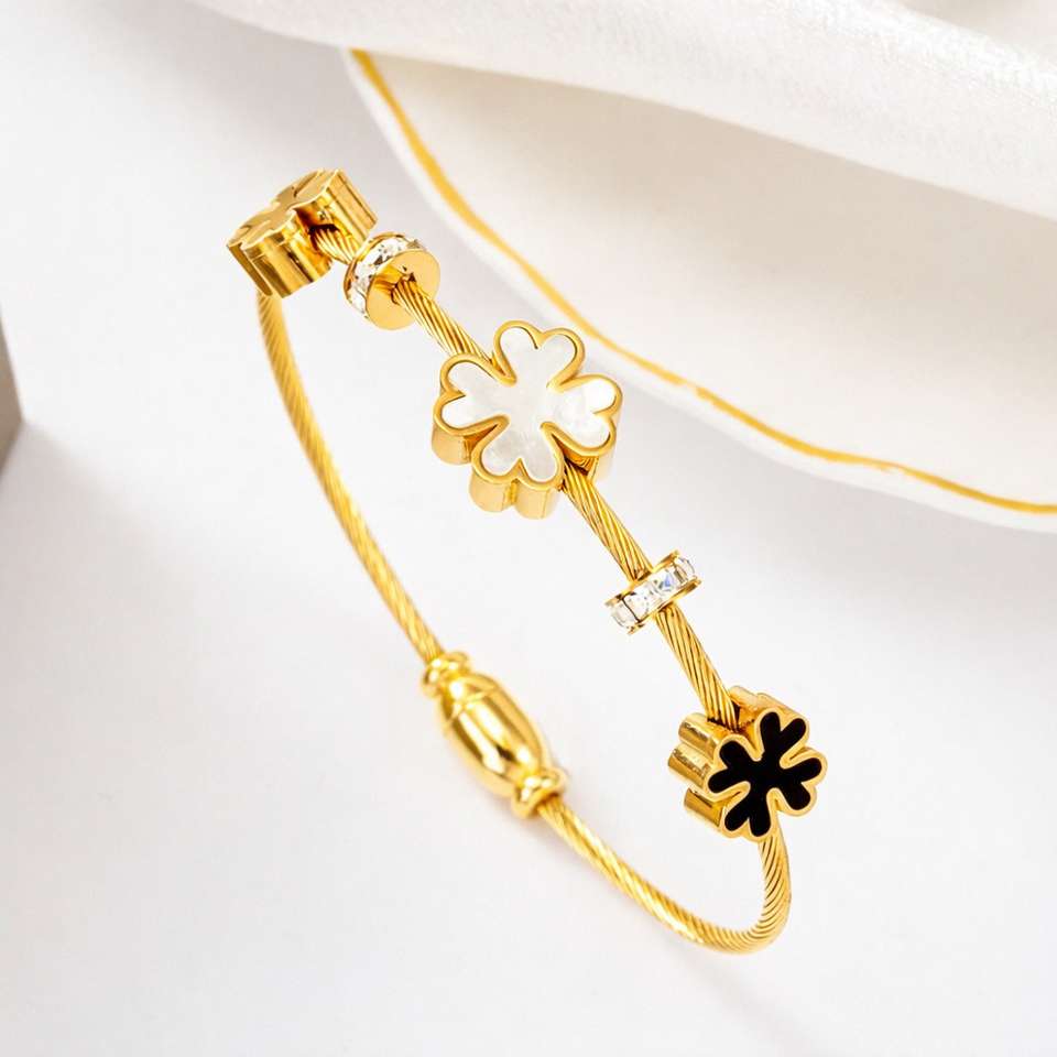 A gold cable bracelet with decorative charms, including two white four-leaf clover charms, a small clear rhinestone accent, and one black four-leaf clover charm, displayed on a soft white background.