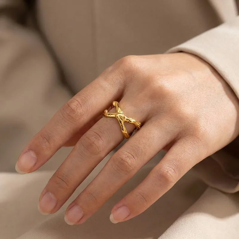 A close-up of a hand wearing a gold ring with an abstract, intertwined organic branch-like design, set against a soft beige background.
