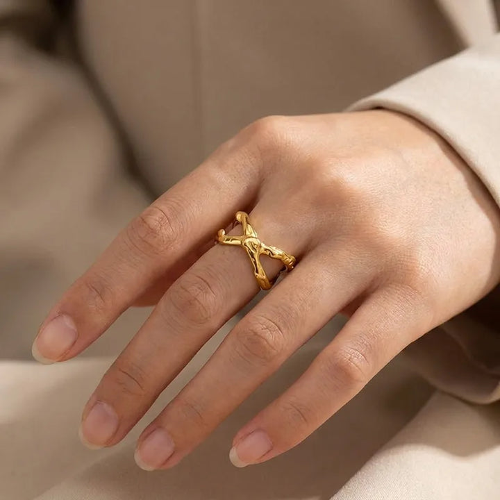 A close-up of a hand wearing a gold ring with an abstract, intertwined organic branch-like design, set against a soft beige background.