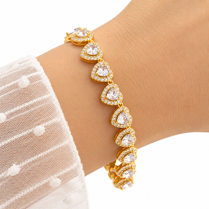 A gold heart-link bracelet with clear heart-shaped crystals and small pavé accents, shown worn on a wrist with a white lace sleeve visible.