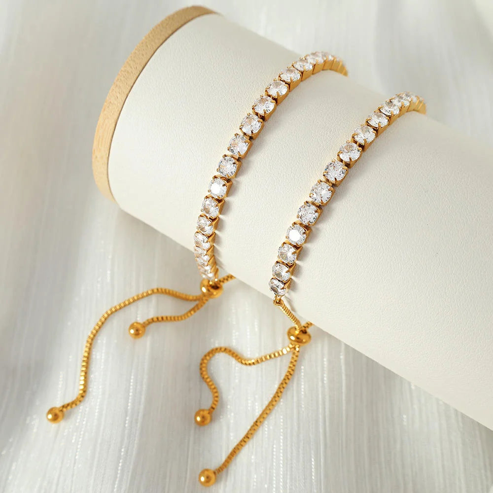 Two gold-tone tennis bracelets with clear rhinestones, draped over a white cylindrical jewelry display stand on a white textured fabric background.